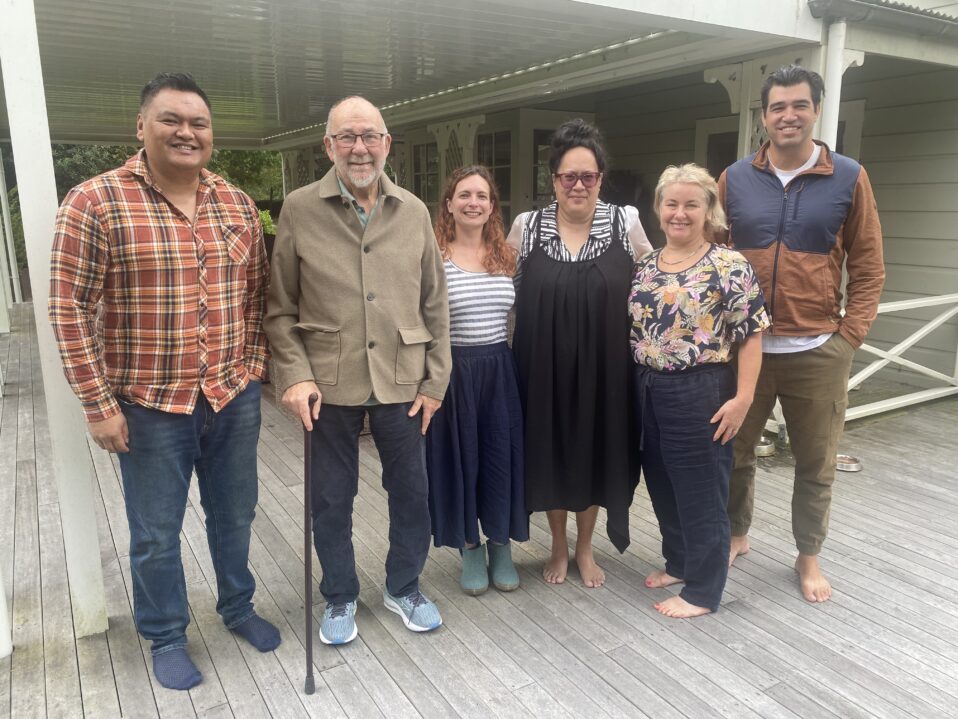 6 people are standing on a porch, three men and three women. From left to right, Patrick Gemmell, CE Tangata Whenua, Tā Kim Workman, CE Tangata Tiriti Katie Bruce, Tangata Whenua Co-chair Maraea Turketo, Tangata Tiriti co-chair Ginnie, and facilitator Haiona Waititi